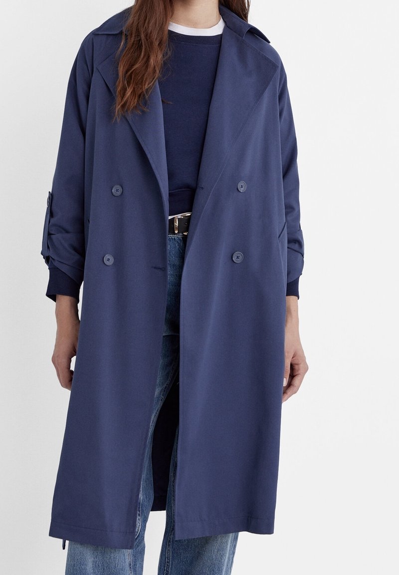 Femme portant un long manteau bleu marine à double boutonnage par-dessus un pull sombre et un jean bleu avec une ceinture noire, debout devant un fond uni.
