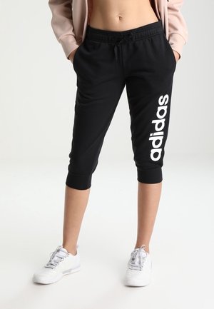 Person iført sorte Adidas capri bukser med hvidt logo på højre ben og hvide sneakers, hænderne i lommerne, stående på en hvid baggrund.