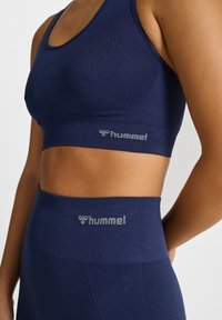 Femme portant un soutien-gorge de sport bleu marine assorti sans coutures et des leggings taille haute, tous deux marqués du logo argenté "hummel".