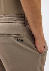 Κοντινό πλάνο του χεριού ενός ατόμου που μπαίνει στην τσέπη μπεζ παντελονιού με ελαστική μέση και μαύρο λογότυπο «BOSS» κοντά στην πίσω τσέπη.