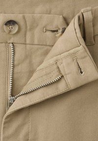 Gros plan d'un pantalon beige avec une fermeture éclair métallique partiellement ouverte et deux boutons visibles sur la ceinture avec des boutonnières.