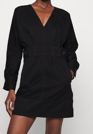 Femme portant une robe mini noire à manches longues avec un col en V, une large ceinture avec boutons, et des poches devant, debout devant un fond uni.