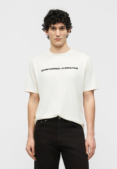 Νεαρός άνδρας με σγουρά σκοτεινά μαλλιά, ντυμένος με λευκό t-shirt Emporio Armani και μαύρο παντελόνι, μπροστά σε απλό φόντο.