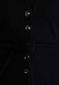 Vestuário de polar preto com cintura amarrada, apresentando botões escuros com um detalhe dourado, design estruturado e textura suave.