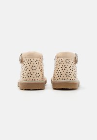Beige perforēti potītes zābaci ar spīdīgu apdari, platu gumijas zoli un dekoratīvām pušķu detaļām aizmugurē.