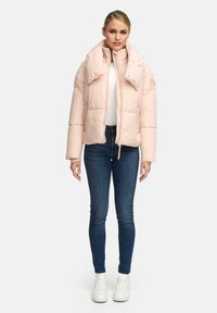Frau, die eine hellrosa Steppjacke über einem weißen Oberteil, dunkelblauen Skinny Jeans und weißen Sneakers trägt, steht vor einem schlichten weißen Hintergrund.