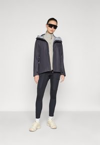 Donkergrijze waterdichte jas met lichtgrijze capuchon, gelaagd over een beige zip-up top en zwarte sportlegging. Het schoeisel bestaat uit lichtgekleurde sneakers.
