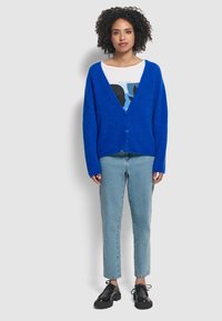 Blauwe donzige cardigan met een diepe V-hals en drie knopen, gedragen over een grafisch wit shirt en gecombineerd met lichtblauwe jeans en zwarte schoenen.