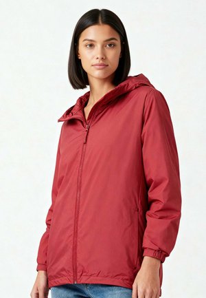 Femme aux cheveux noirs raides portant une veste à capuche rouge zippée et un jean bleu, debout devant un fond clair uni.