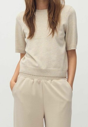 Kvinde iført lysebeige kortærmet striktop og matchende elastikbuks med hænderne i lommerne mod ensfarvet baggrund.