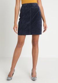 Marinblå mockaskjol med en elegant penningform, som har hög midja och subtila sömnadsdetaljer. Matchas med svarta och vita randiga klackar.