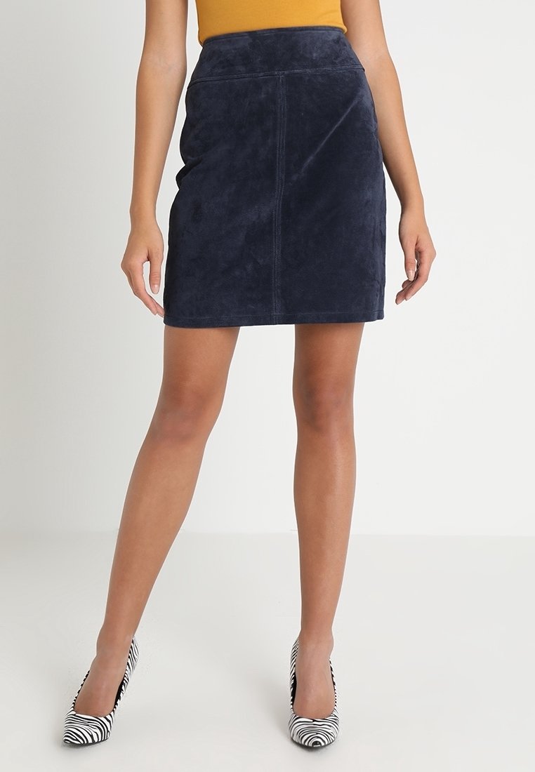 Marinblå mockaskjol med en elegant penningform, som har hög midja och subtila sömnadsdetaljer. Matchas med svarta och vita randiga klackar.