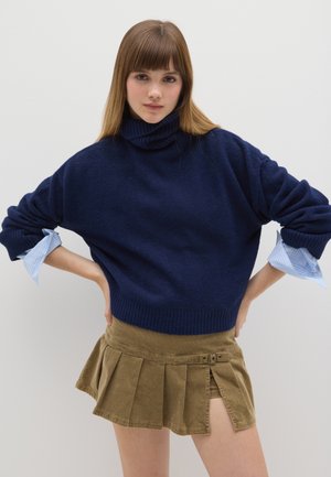 Maglione a collo alto blu navy indossato sopra una camicia a righe blu chiaro, abbinato a una minigonna plissettata marrone con fibbia laterale.