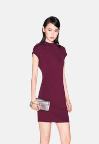 Abito fitted di color bordeaux con scollo alto e maniche a cappuccio. La modella tiene una pochette argentata con tracolla, posata su uno sfondo neutro.