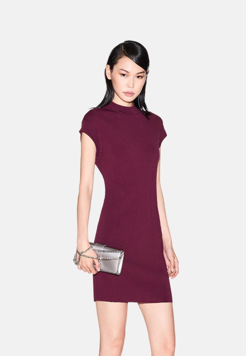 Abito fitted di color bordeaux con scollo alto e maniche a cappuccio. La modella tiene una pochette argentata con tracolla, posata su uno sfondo neutro.