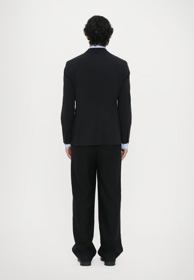 Schwarze strukturierte Anzugjacke mit geradem Rücken und langen Ärmeln, kombiniert mit weit geschnittenen schwarzen Hosen und sichtbaren hellblauen Manschetten.