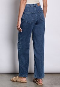 Jeans taille haute, à jambes larges en denim bleu foncé avec poches arrière, lignes droites et une étiquette en cuir beige au niveau de la taille.