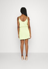Vestido mini de cetim verde com alças finas, apresentando uma parte de trás baixa e uma cintura ajustada. Sandálias de salto bloco transparentes completam o visual.