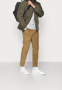 Olivgrön puffjacka, ljusgrå stickad tröja, tan-färgade kordbyxor och vita sneakers, med en svart ryggsäck som bärs på ena axeln.