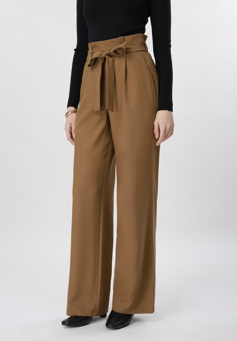 Femme portant un pantalon large marron taille haute avec une ceinture nouée, un haut noir à manches longues, des chaussures noires et un bracelet au poignet gauche.