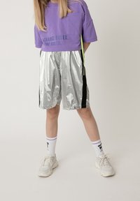 Pantaloni corti riflettenti argento con pannelli laterali neri, abbinati a un top corto viola con taschino e testo stampato. Sneakers bianche e calzini a righe.