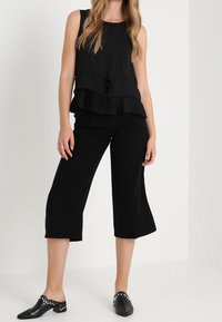 Svart ärmlös topp med lager av veck och knytDetalj, kombinerad med svarta culotter. Modellen bär svarta mules med nitar.