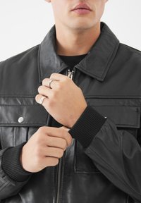 Mann in schwarzer Lederjacke mit gerippten Bündchen, der den Ärmel anpasst und zwei silberne Ringe an der linken Hand vor weißem Hintergrund zeigt.
