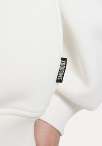 Felpa bianca con polsini a costine, caratterizzata da un'etichetta nera sulla manica. Il materiale sembra morbido e strutturato. Design slim fit.