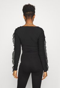 Svart långärmad topp med volangdetaljer av genomskinligt material på ärmarna. Åtsittande design med mjuk textur, rund halsringning och veckad detaljering.