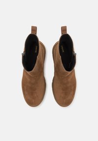 Bruna mockaskinnsankleboots med en slät yta, med en sidodragkedja och en ögla för att dra upp dem, placerade på en ljus bakgrund.