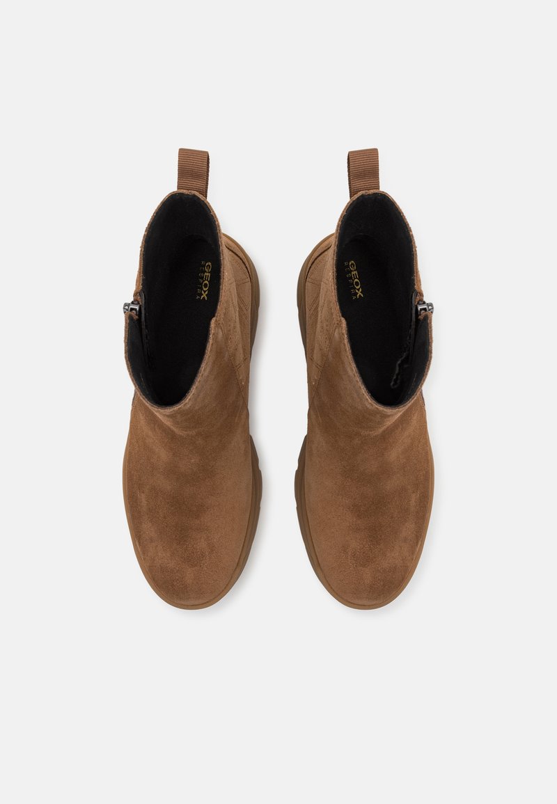 Botas de ante marrón hasta el tobillo con una textura suave, que cuentan con una cremallera lateral y una lengüeta de tira, ubicadas sobre un fondo claro.