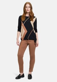 Schwarzes, tanfarbenes und braunes Langarmoberteil mit geometrischem Design und Verzierungen, kombiniert mit braunen, körpernahen Hosen und schwarzen Slippern.