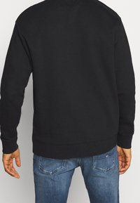 Sudadera de algodón negra con cuello redondo y puños acanalados, vista desde la parte posterior. Combinada con jeans de mezclilla azules con bolsillos.