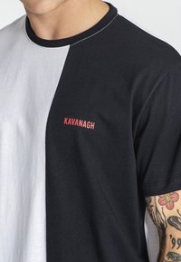 Čiernobiely tričko s farebnými blokmi vyrobené z bavlny, s okrúhlym výstrihom a nápisom "Kavanagh" v červenom písme na hrudi.