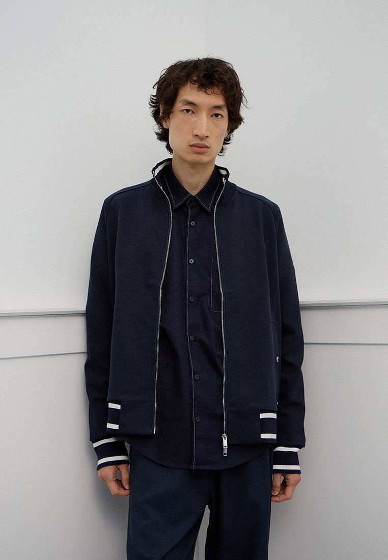 Granatowa kurtka zapinana na zamek z wysokim kołnierzem, z białymi paskami na mankietach i u dołu, noszona na dopasowanej ciemnej koszuli, z detalem kieszeni.