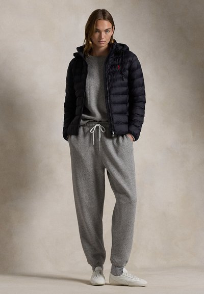 Mladá žena nosící černou prošívanou bundu přes šedou mikinu a tepláky, stojí s rukama v kapsách na neutrálním pozadí.