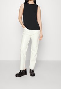 Schwarzes ärmelloses Top kombiniert mit cremefarbenen, taillierten Hosen und glänzenden schwarzen Wedge-Schuhen. Glatter Stoff mit klaren Linien, ohne Muster.