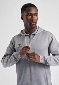 Grijze zip-up hoodie met een trekkoord in de capuchon, gemaakt van gestructureerde stof. Bevat een logo op de borst en contrasterende stikseldetails.