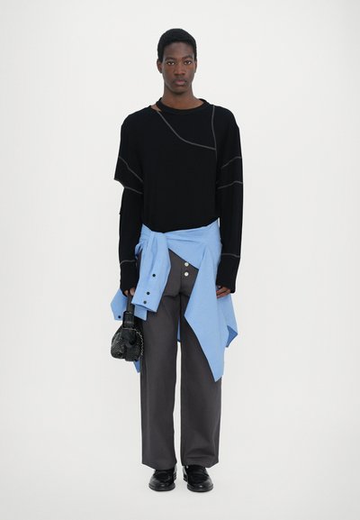 Black long-sleeve top with beige stitching, light blue shirt tied at the waist, gray wide-leg pants, and black shoes. Holds a small black bag.