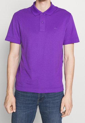 Homme portant un polo violet uni à manches courtes avec un col et deux boutons, associé à un jean bleu, debout devant un fond neutre.
