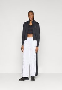 Černá zipová bunda s vysokým límcem, spárovaná s černým crop topem a bílými kalhotami se stranovým logem. Model nosí černou obuv.