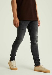 Černé džíny z denimové látky s opotřebovanými detaily, slim fit, doplněné béžovými teniskami s texturovanou podrážkou a šněrováním, na neutrálním pozadí.