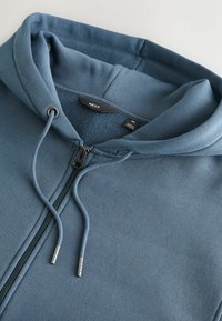 Sweat à capuche bleu moyen avec fermeture éclair, cordons de serrage, doublure en polaire, étiquettes indiquant la marque "Next" et la taille "M".