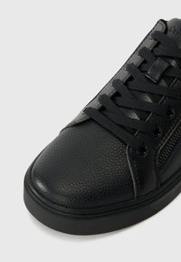 Close-up af sort struktureret læder-sneaker med sorte snørebånd, metaløjer og synlig sidelås på hvid baggrund.