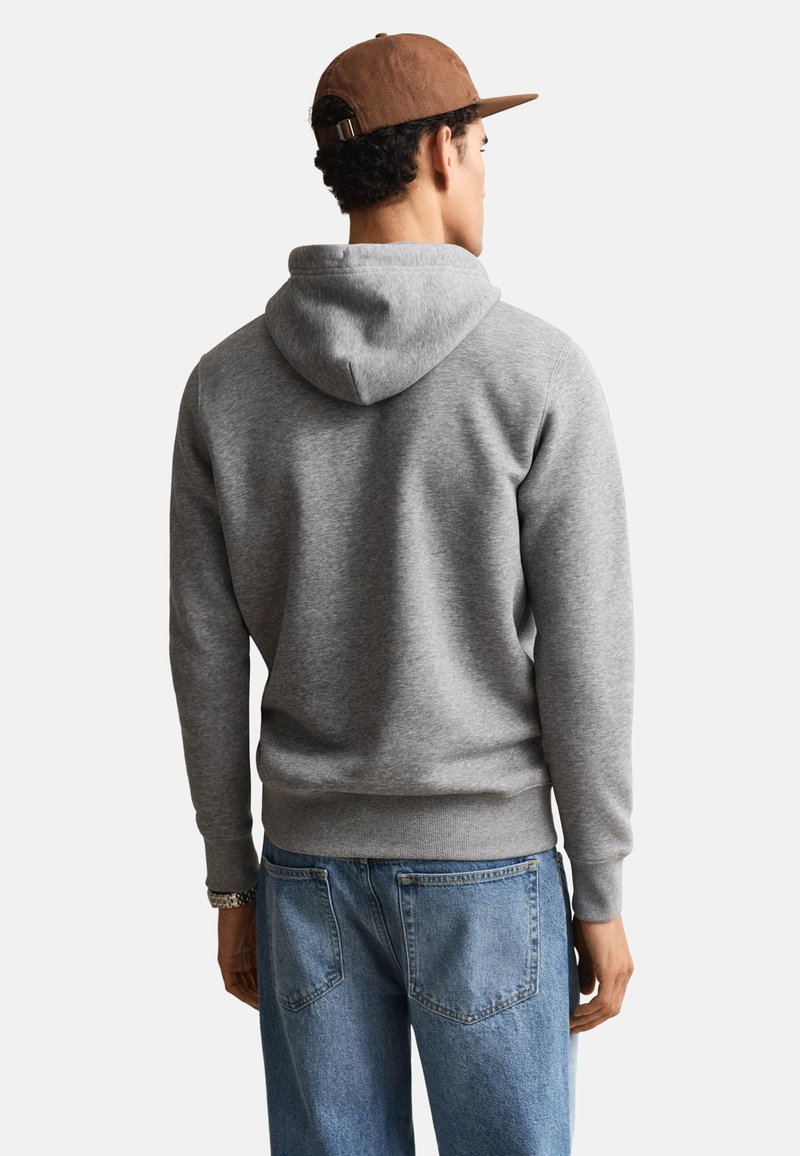 Hombre que lleva una gorra marrón, sudadera gris con capucha y vaqueros azules, mirando hacia atrás con un fondo blanco liso.