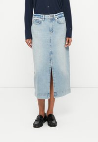Saia midi de ganga azul clara com uma racha frontal, cintura alta e costura visível. Usada com sapatos pretos de entrar para contraste.