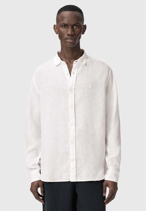 Mand, der står fremadrettet iført en hvid linnedskjorte med lange ærmer og knapper samt mørke bukser foran en ensfarvet baggrund.