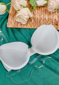 Biały stanik z wyściółką, gładkim materiałem, fiszbinami i małym detalem w postaci kokardki. Przezroczyste regulowane ramiączka leżą na zielonym tle.