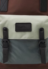 Hnědý a zelený batoh s texturovaným horním klapkou, černými koženými detaily a dvěma přezkami na přední kapse.