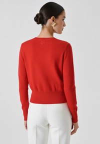 Eine Frau mit dunklem Haar, das zu einem Dutt gebunden ist, trägt ein rotes Langarmoberteil und weiße Hosen. Sie wird von hinten vor einem schlichten grauen Hintergrund gezeigt.